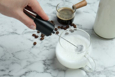 Woman whisking milk in cup with mini mixer (milk frother) at white marble table, closeup