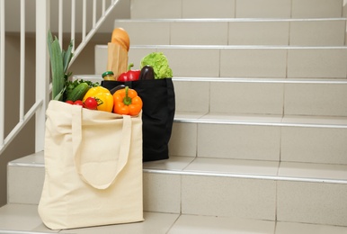 Photo of Tote bags with vegetables and other products on stairs indoors. Space for text