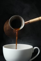 Turkish coffee. Pouring brewed beverage from cezve into cup against black background