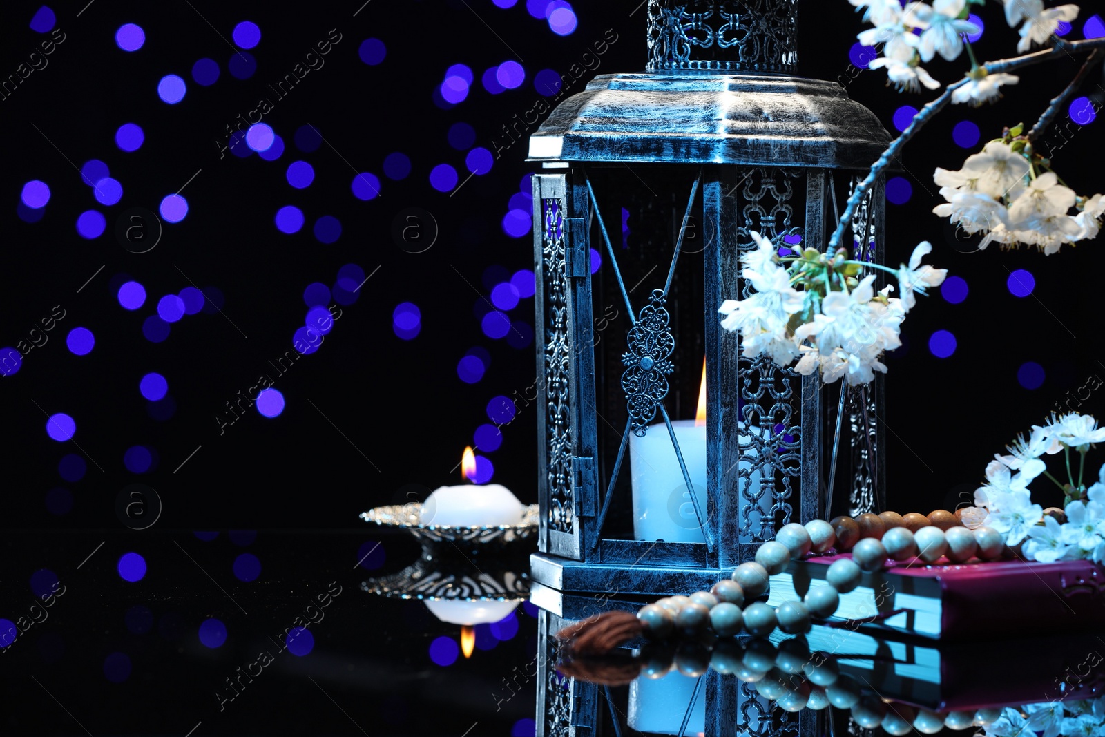 Photo of Arabic lantern, Quran, misbaha, burning candle and flowers on mirror surface against blurred lights at night. Space for text