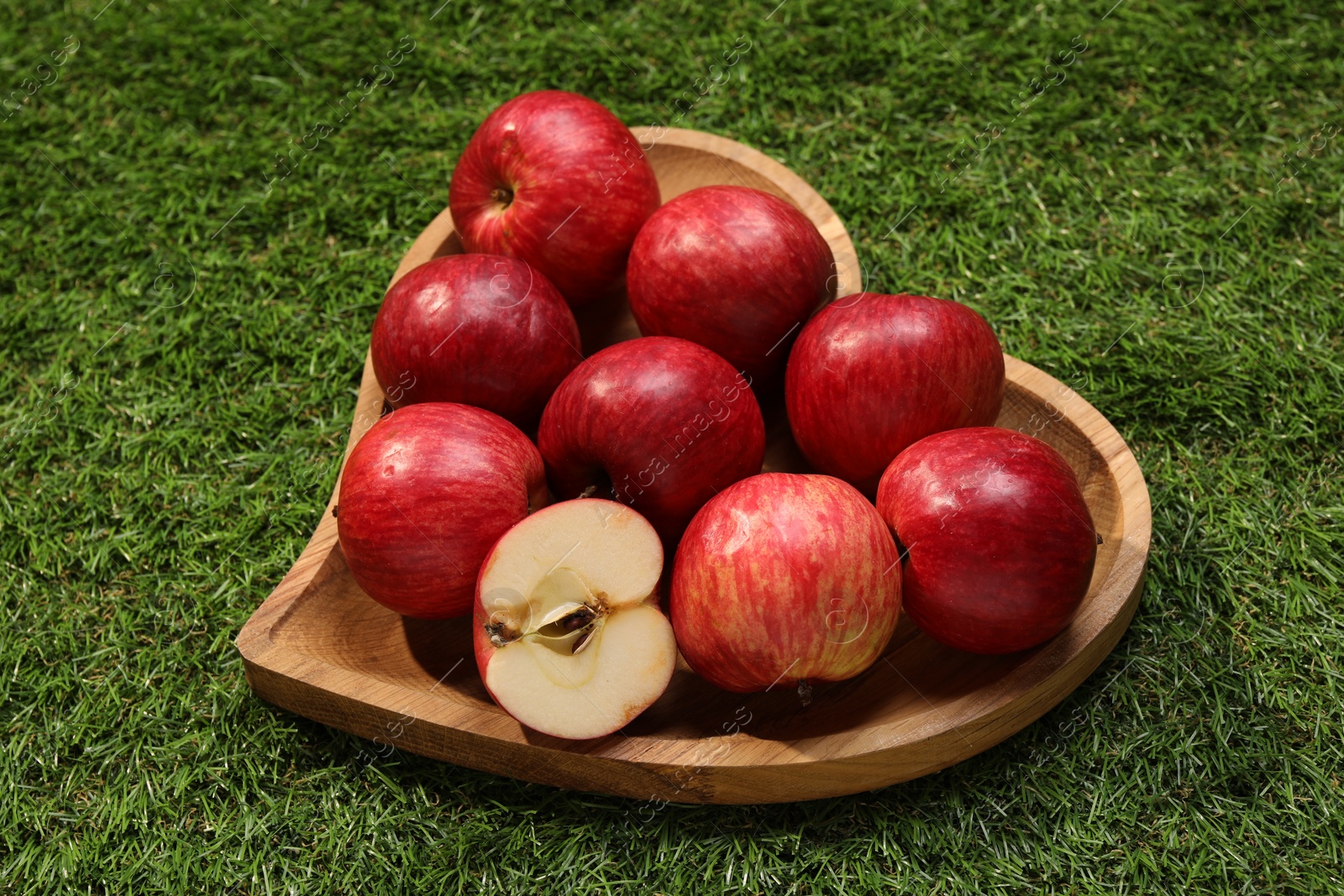 Photo of Wooden plate in shape of heart with red apples on green grass