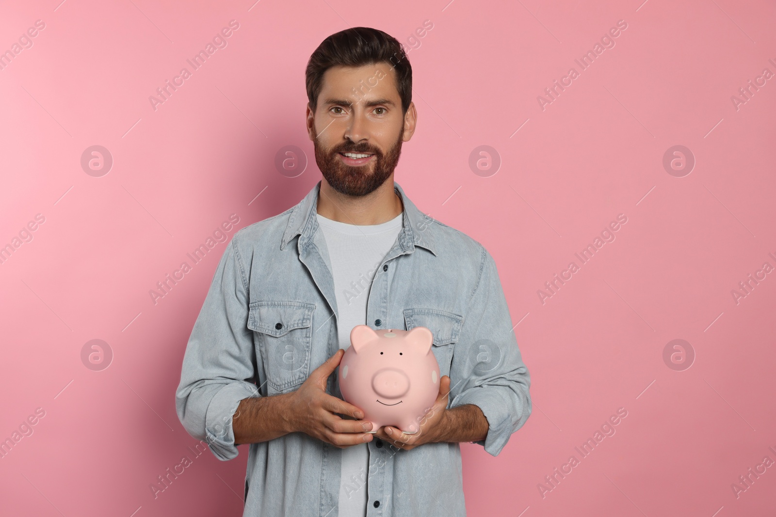 Photo of Happy man with ceramic piggy bank on pale pink background