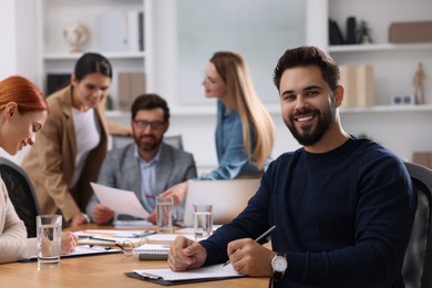 Team of employees working together in office. Happy man with pen and clipboard at table indoors