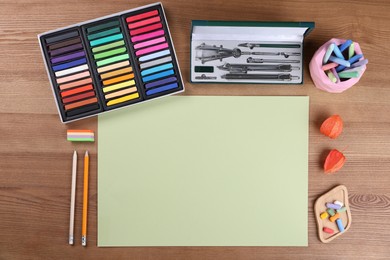 Photo of Blank sheet of paper, colorful chalk pastels and other drawing tools on wooden table, flat lay. Modern artist's workplace