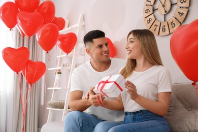 Man presenting gift to his girlfriend in room decorated with heart shaped balloons. Valentine's day celebration