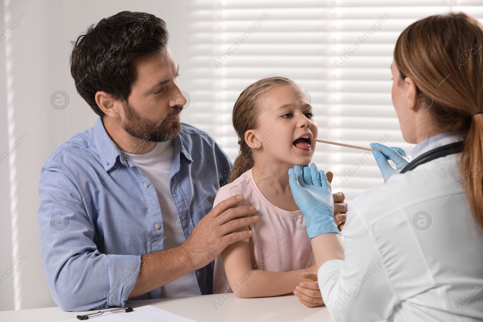 Photo of Doctor examining girl`s oral cavity with tongue depressor near her father indoors