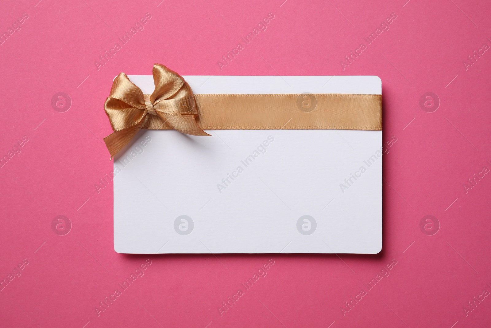 Photo of Blank gift card with golden bow on pink background, top view. Space for text