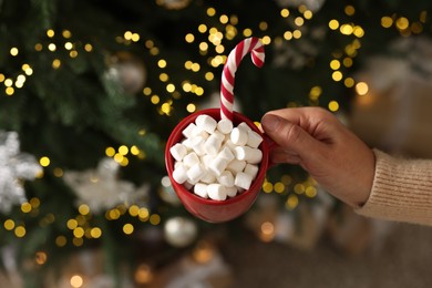 Photo of Woman holding cup of delicious Christmas cocoa with marshmallows and candy cane near fir tree, above view