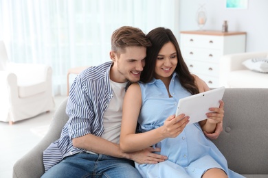Photo of Young man and his pregnant wife using tablet computer on sofa at home