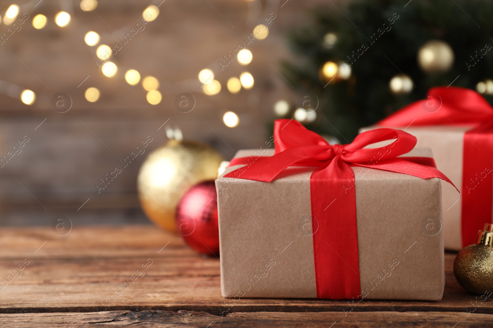 Photo of Christmas gift box with red ribbon and golden bauble on wooden table against blurred lights, space for text