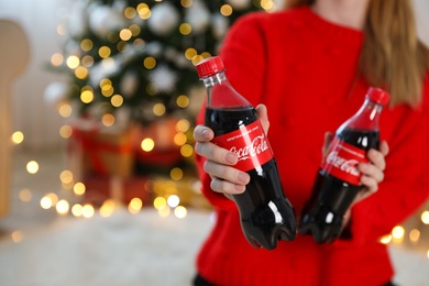 Photo of MYKOLAIV, UKRAINE - January 01, 2021: Woman with bottles of Coca-Cola against blurred Christmas tree indoors, closeup