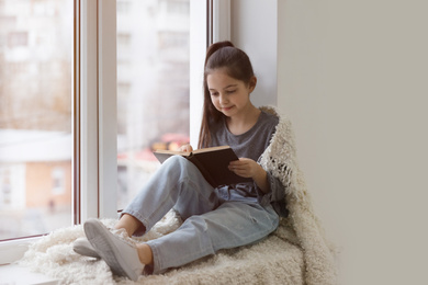 Cute little girl reading book near window at home