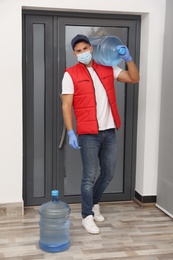 Photo of Courier in face mask with bottles of cooler water in entryway. Delivery during coronavirus quarantine