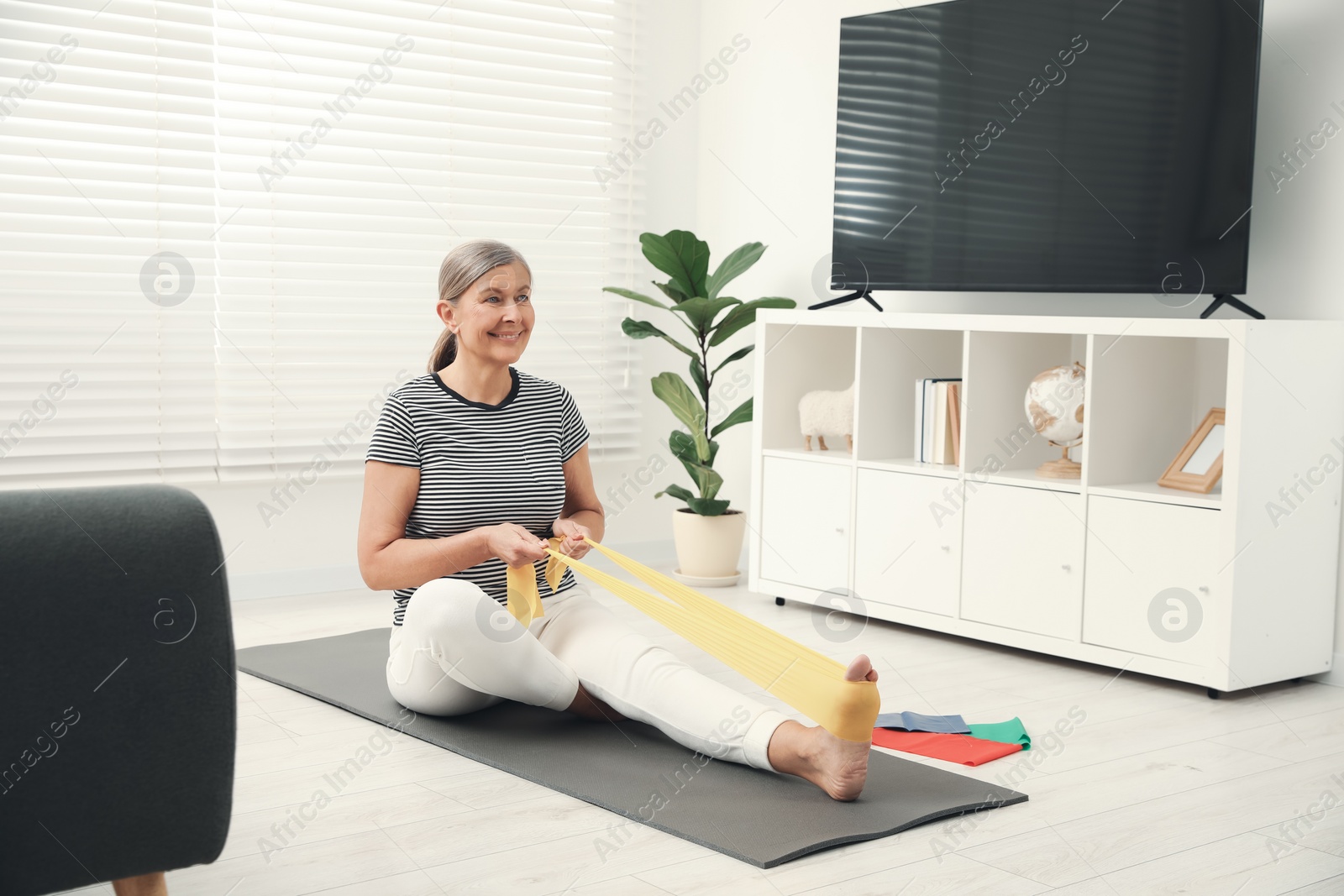 Photo of Senior woman doing exercise with fitness elastic band on mat at home