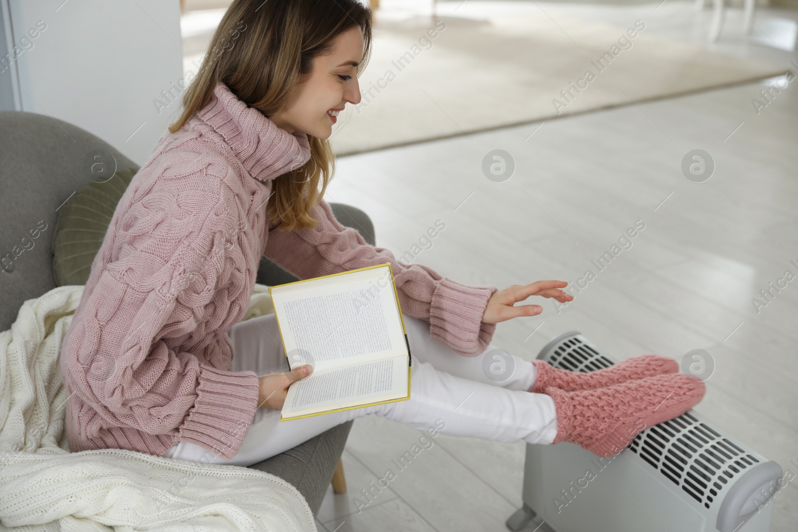 Photo of Happy young woman with book warming feet on electric heater at home