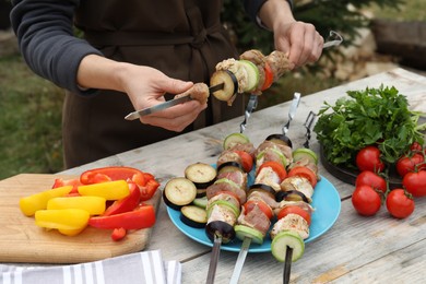 Photo of Woman stringing marinated meat and vegetables on skewer at wooden table outdoors, closeup