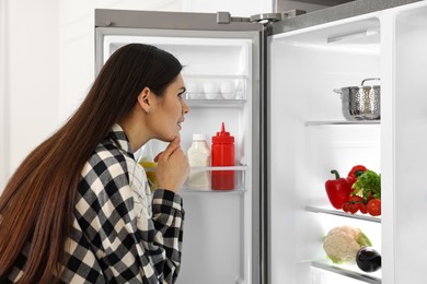 Thoughtful young woman near modern refrigerator indoors