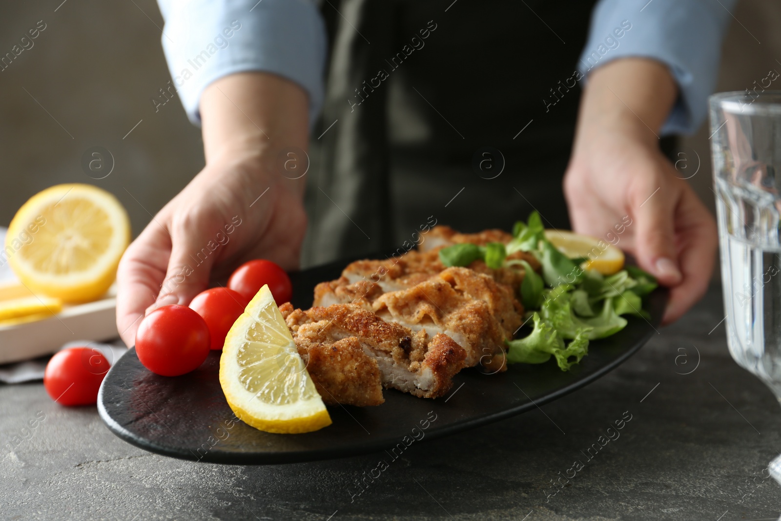 Photo of Woman holding tasty schnitzel with tomatoes, greens and lemon at grey textured table, closeup