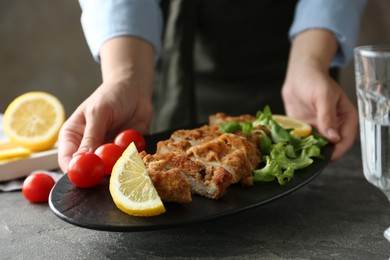 Woman holding tasty schnitzel with tomatoes, greens and lemon at grey textured table, closeup