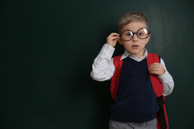 Photo of Cute little child wearing glasses near chalkboard, space for text. First time at school