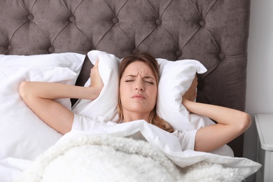 Photo of Young woman covering ears with pillow while trying to sleep in bed at home. Early morning