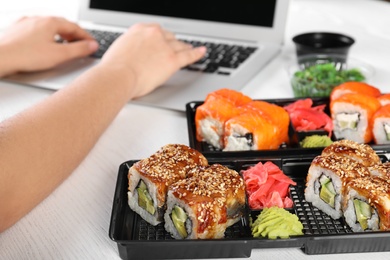 Boxes with different sushi rolls and blurred woman using laptop on background. Food delivery