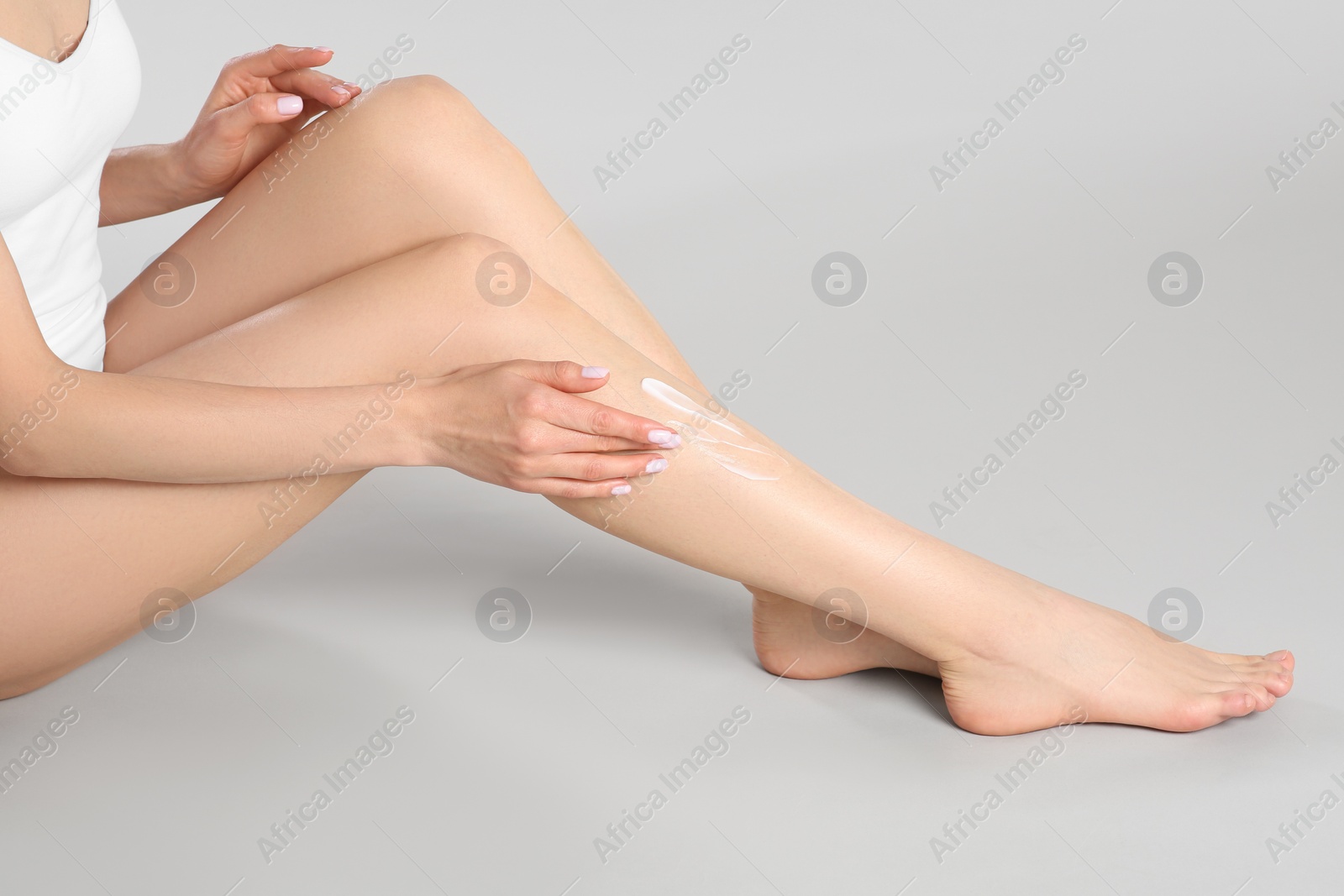 Photo of Woman applying body cream onto her smooth legs on light grey background, closeup