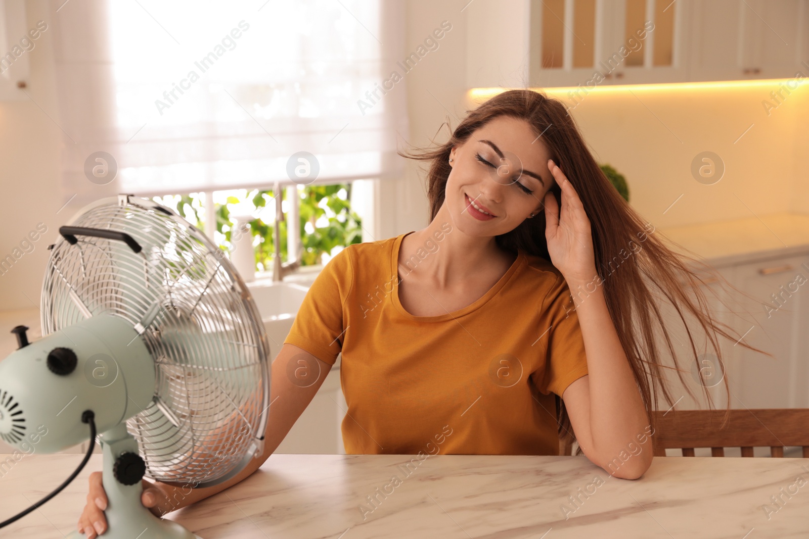 Photo of Woman enjoying air flow from fan at table in kitchen. Summer heat