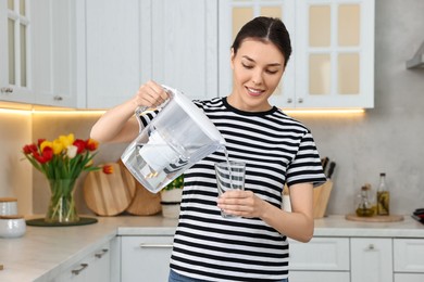 Photo of Woman pouring water from filter jug into glass in kitchen