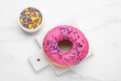 Photo of Sweet glazed donut decorated with sprinkles on white marble table, flat lay. Tasty confectionery