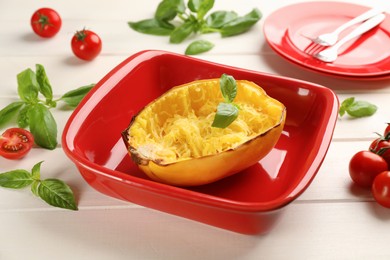 Photo of Half of cooked spaghetti squash with basil in baking dish and tomatoes on white wooden table