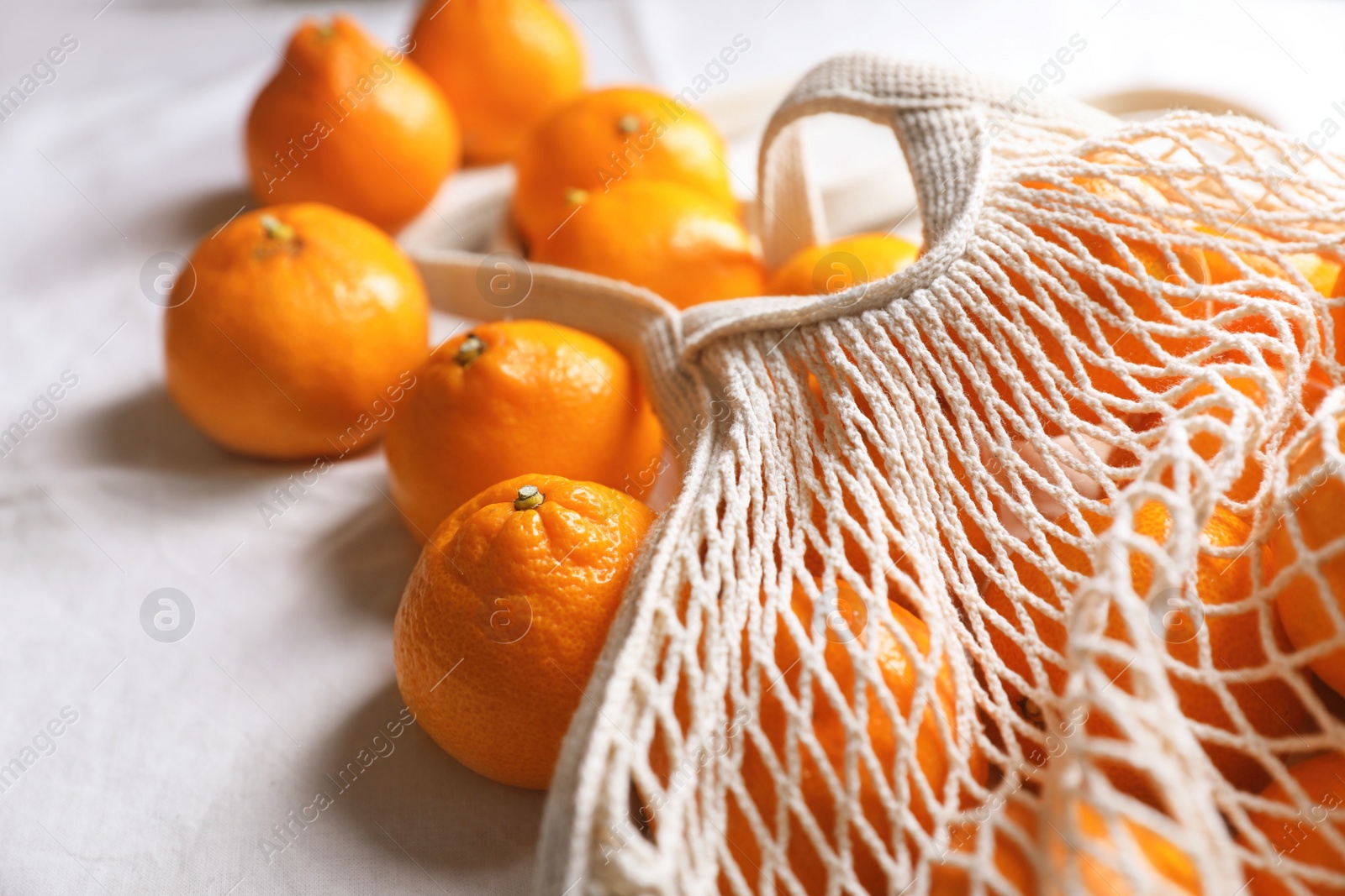 Photo of Net bag with many fresh ripe tangerines on white cloth, closeup