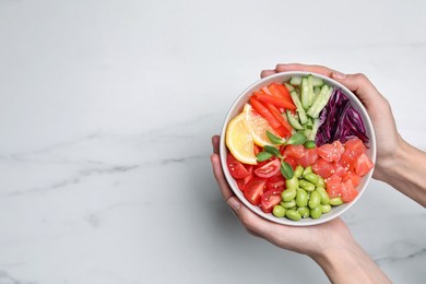 Woman holding poke bowl with salmon, edamame beans and vegetables at white marble table, top view. Space for text
