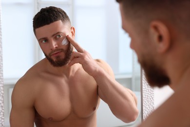 Photo of Handsome man applying cream onto his face near mirror in bathroom