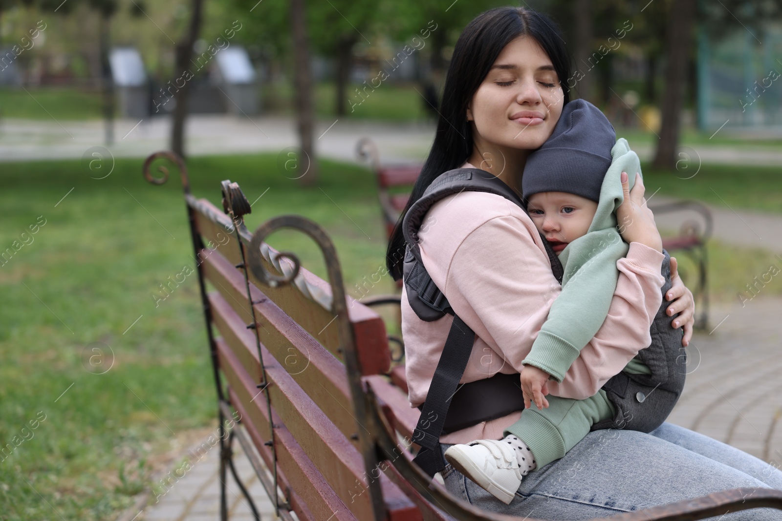 Photo of Mother holding her child in sling (baby carrier) on bench in park