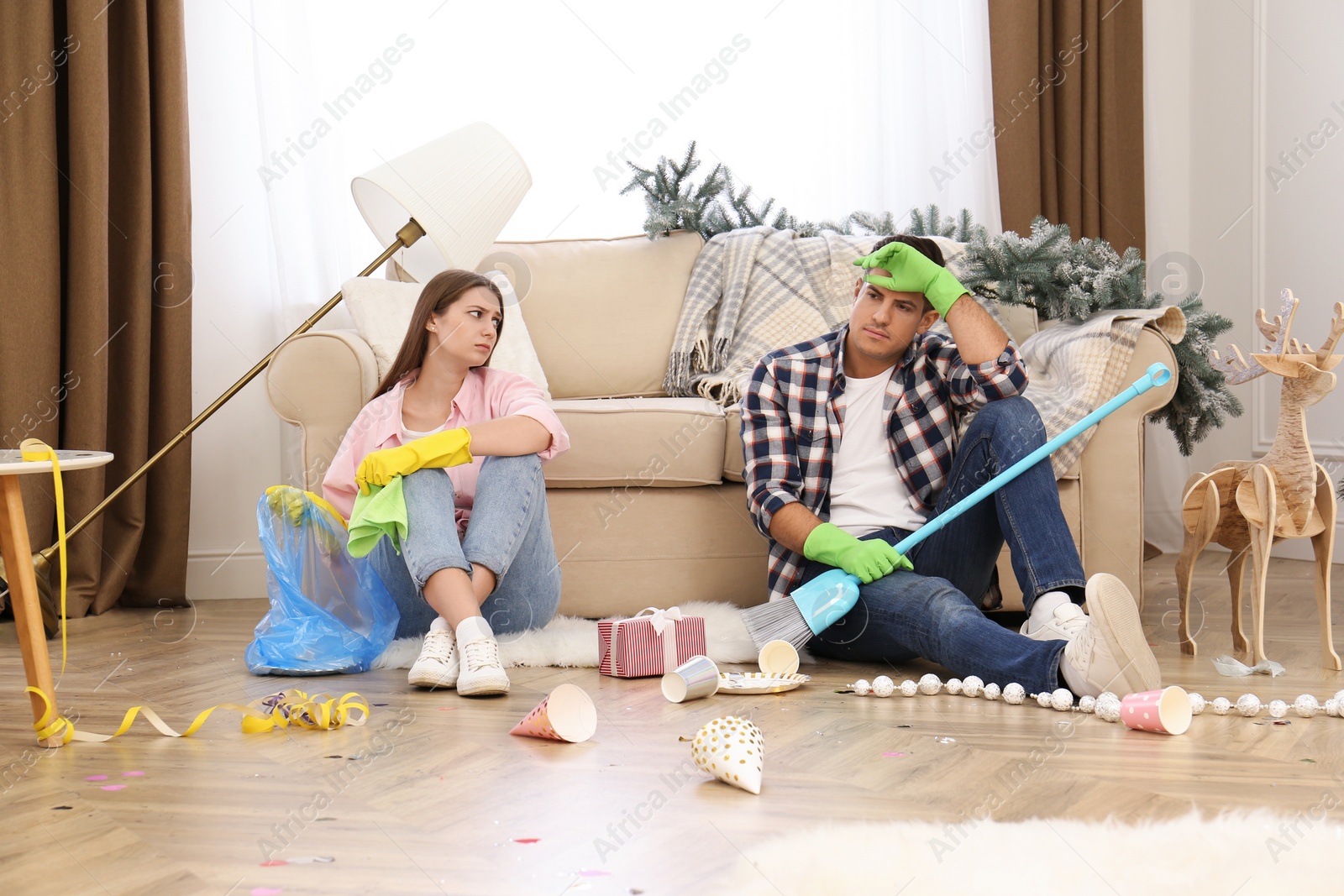 Photo of Tired couple sitting in messy room while cleaning after New Year party