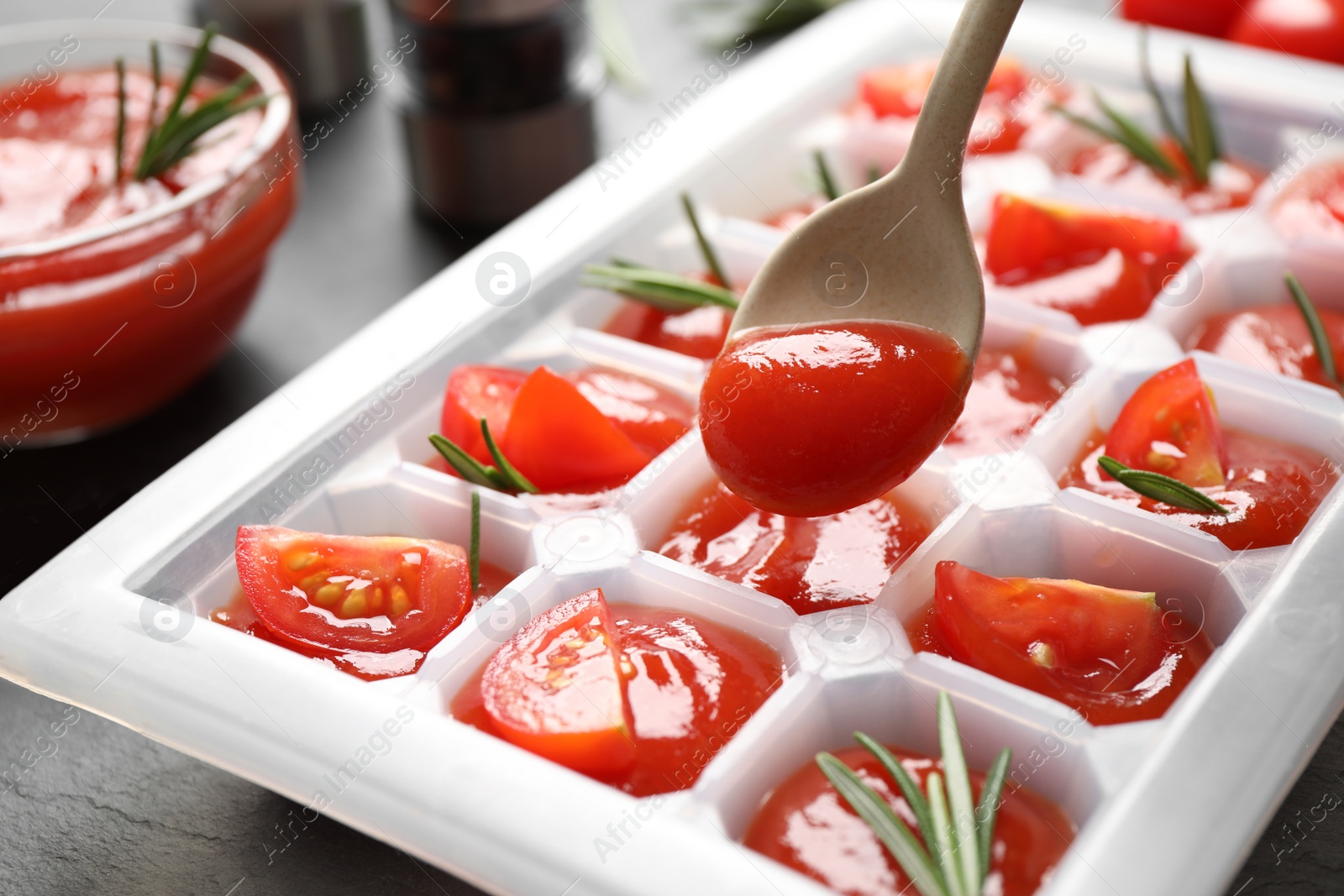 Photo of Pouring sauce into ice cube tray with tomatoes and rosemary on grey table, closeup