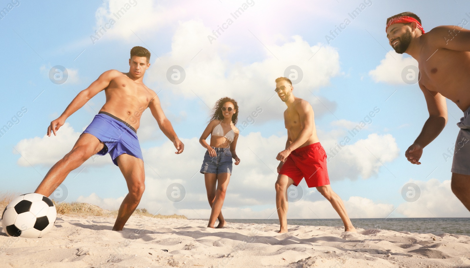 Image of Happy friends playing football on beach during sunny day, low angle view