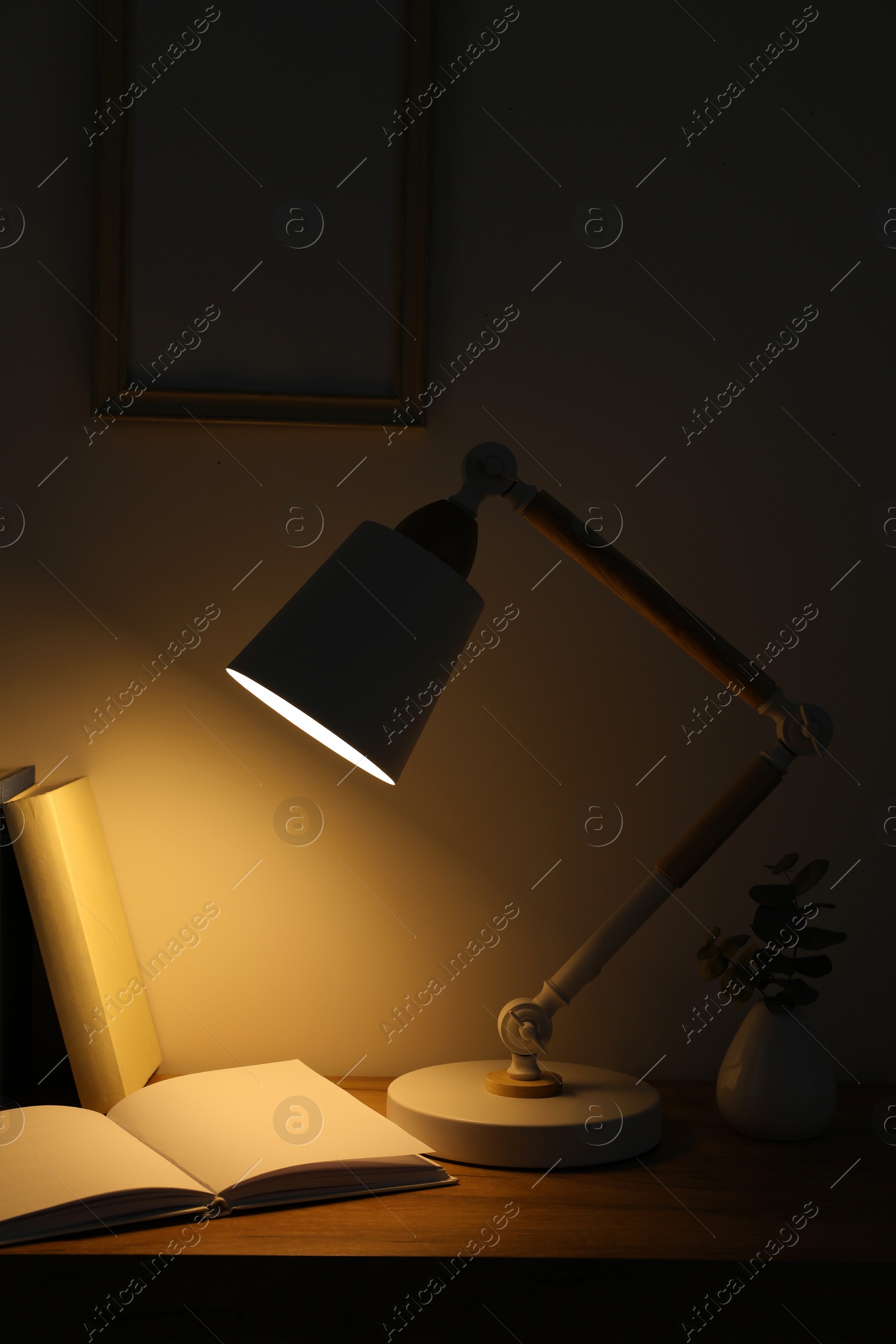 Photo of Stylish modern desk lamp, books and plant on wooden table in dark room