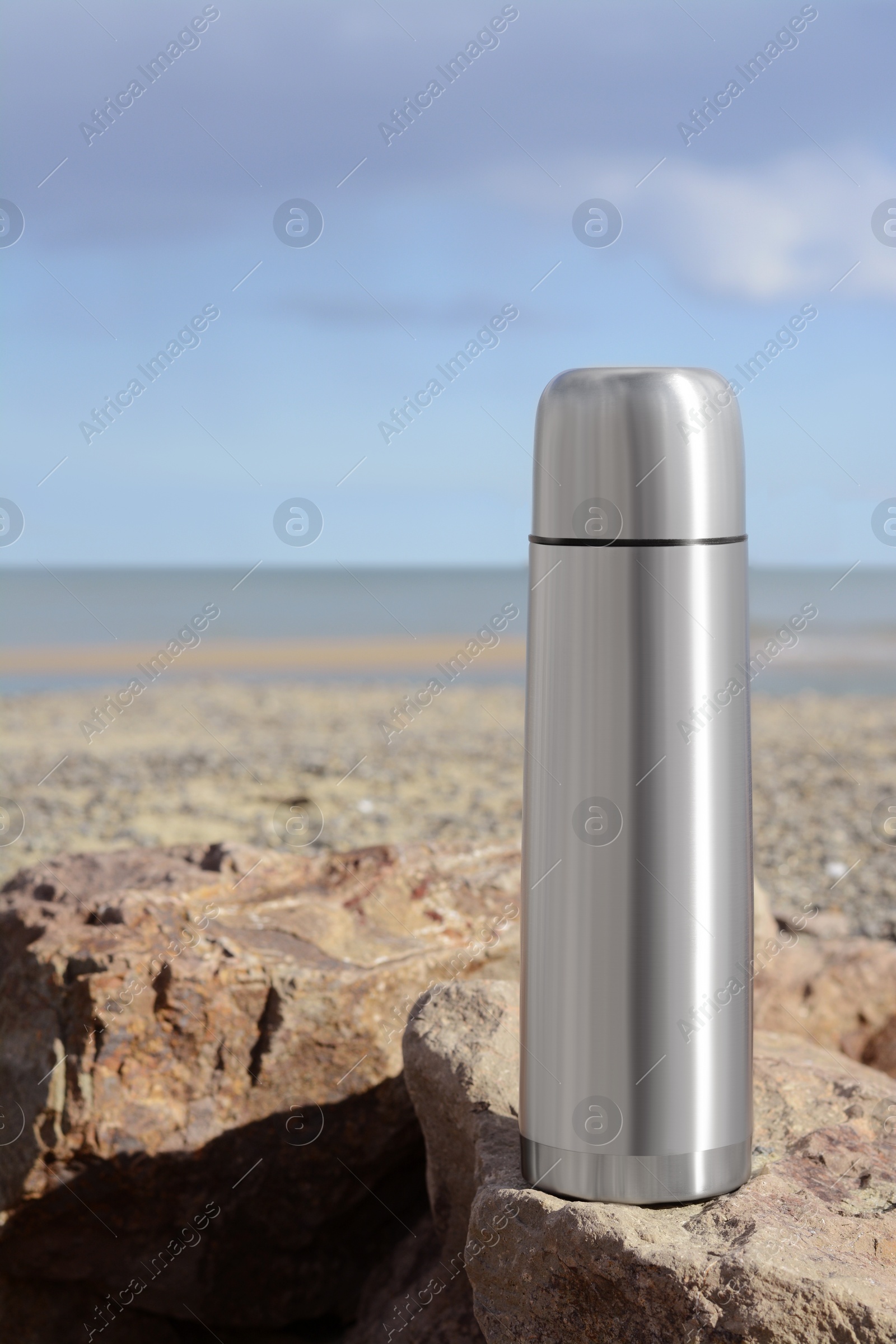 Photo of Metallic thermos with hot drink on stone near sea, space for text