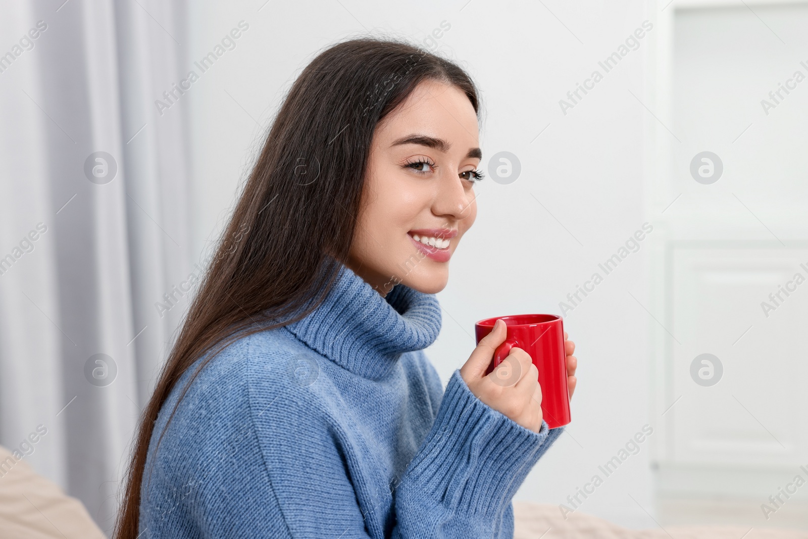 Photo of Happy young woman with red ceramic mug on bed at home, space for text