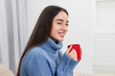 Happy young woman with red ceramic mug on bed at home, space for text