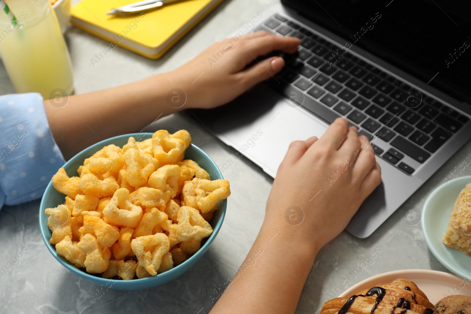 Photo of Bad eating habits. Woman using laptop surrounded by different snacks at light grey marble table, closeup