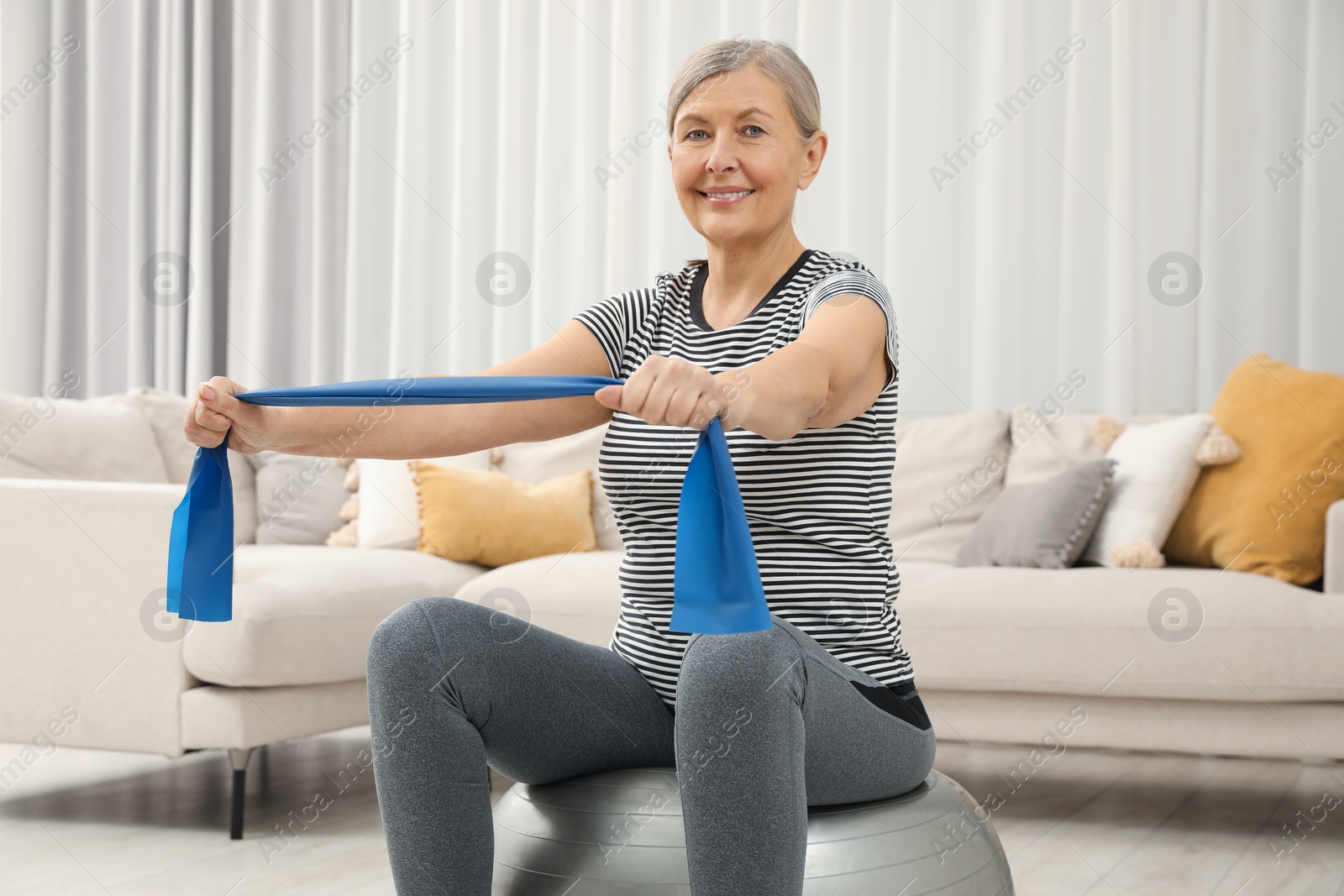 Photo of Senior woman doing exercise with elastic resistance band on fitness ball at home