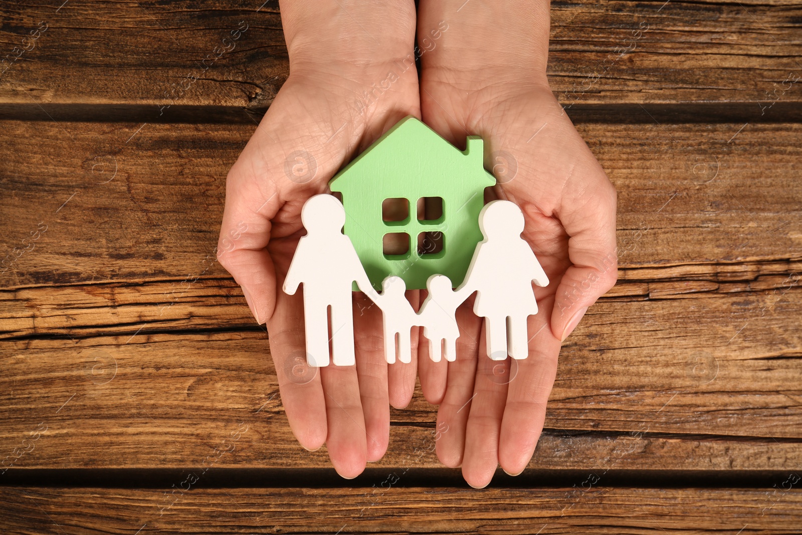 Photo of Woman holding figures of family and green house in hands on wooden background, top view