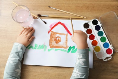 Cute little girl drawing house with brush at wooden table, top view. Child`s art