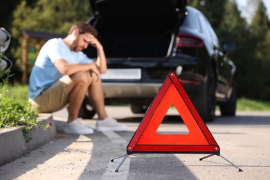 Photo of Man sitting near broken car on roadside outdoors, focus on warning triangle