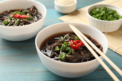 Photo of Tasty soup with buckwheat noodles (soba), chili pepper, green onion in bowl and chopsticks on light blue wooden table, closeup