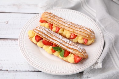 Photo of Delicious eclairs filled with cream, strawberries and mint on white wooden table