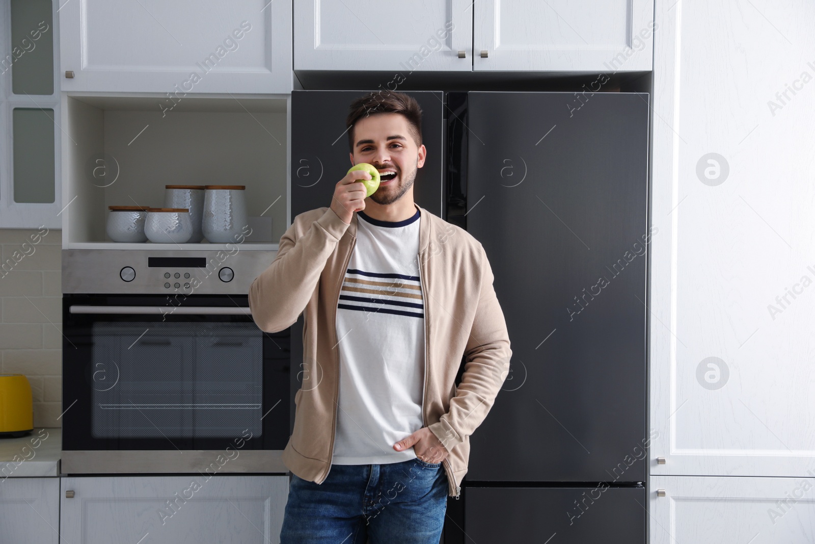 Photo of Young man eating apple near refrigerator in kitchen